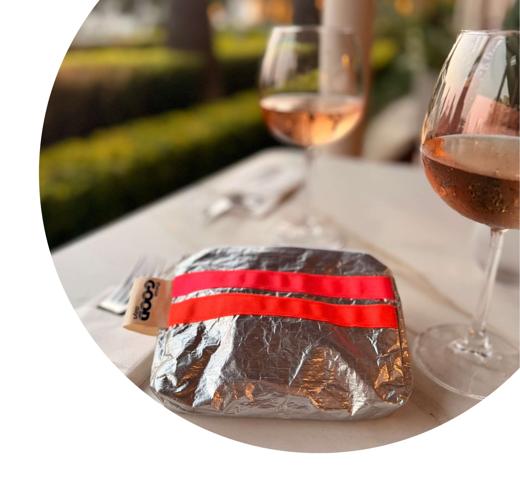 A silver  pouch with a bright pink and orange ribbons sits on a white table, with two wine glasses in the background and a blurred outdoor setting. A small tag with the word 'these GOOD days' is visible.